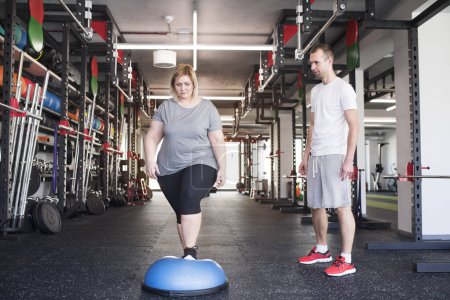 Overweight woman exercising on balance ball in gym. Personal trainer couching and helping her.