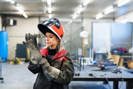 A beautiful blonde woman works as a welder in workshop, operating welding machine, wearing protective clothing and a welding mask.