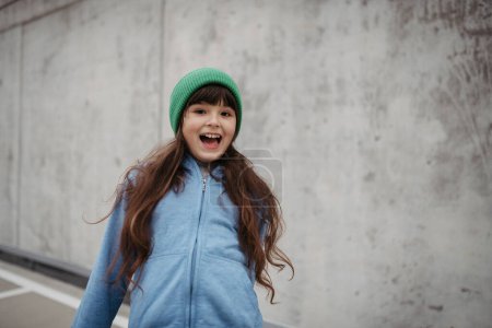 Außenporträt eines süßen jungen Mädchens mit Hut, lachend. Mädchen mit langen Haaren steht auf Spielplatz in der Stadt.