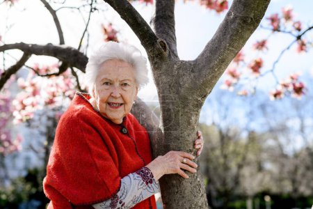 Foto de Retrato de una hermosa anciana parada junto al árbol de magnolia, en el parque, disfrutando de un momento de relax. Abuela disfrutando de clima cálido primavera. - Imagen libre de derechos