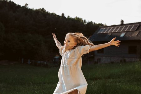 Téléchargez les photos : Petite fille devant la maison avec des panneaux solaires sur le toit, sautant, dansant et s'amusant. Concept de ressources renouvelables, durabilité et énergie verte. - en image libre de droit