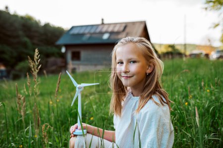 Foto de Chica con modelo de turbina eólica, de pie en el medio de la pradera, casa con paneles solares detrás. Concepto de recursos renovables, sostenibilidad y energía verde. - Imagen libre de derechos