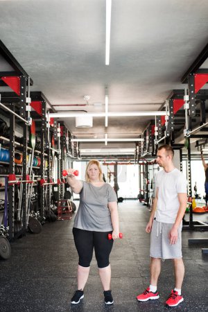 Foto de Overweight woman exercising in gym, using dumbbells. Personal trainer couching her and helping her - Imagen libre de derechos
