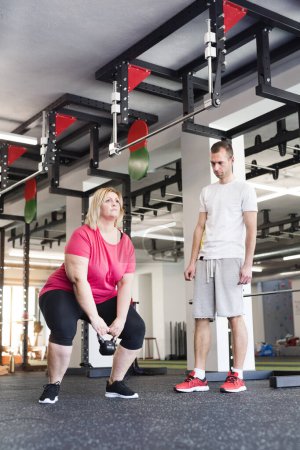 Foto de Mujer con sobrepeso haciendo ejercicio en el gimnasio, usando kettlebell. Entrenador personal que la acaricia y la ayuda - Imagen libre de derechos