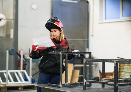 A beautiful blonde woman works as a welder in workshop, operating welding machine, wearing protective clothing and a welding mask.
