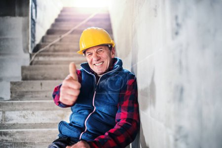Smiling mature man wearing hard hat on construction site, sitting on concrete stairs. Thumbs up.