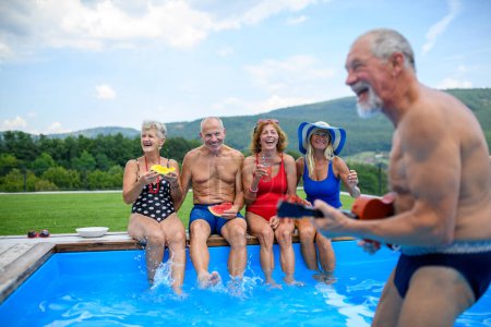 Foto de Grupo de jubilados alegres sentados en el borde de la piscina con las piernas en el agua, comiendo sandía, riendo. Ancianos amigos pasar el día caliente en la piscina, tomar el sol. - Imagen libre de derechos