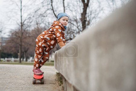 Linda niña jugando al aire libre en el patio de recreo con monopatín. Chica en traje de cuerpo softshell pasar tiempo en el parque de la ciudad.