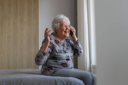 Senior woman at home, phone calling with family member, sitting on bed. Older woman living alone, enjoying peaceful weekend day.