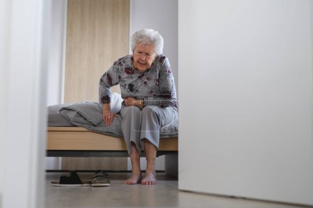 Femme âgée essayant de se lever du lit, ressentant de la douleur, tenant l'abdomen.