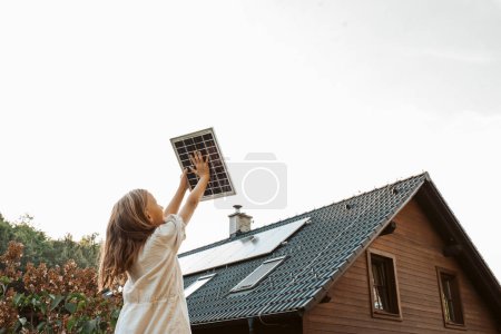 Photo for Little girl with model of solar panel, standing in the middle of meadow, house with solar panels behind. Concept of renewable resources, sustainability and green energy. - Royalty Free Image