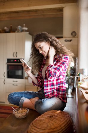 University student sitting on kitchen counter scrolling on smartphone with stiff neck.