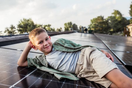 Boy lying on roof with solar panels, looking at camera, smiling. Sustainable future for next generation concept.