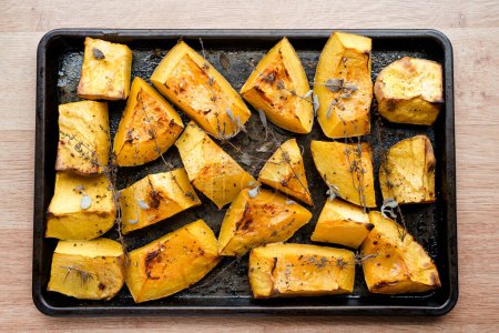 Pieces of organic oven roasted butternut squash on a black baking sheet pan with fresh thyme and sage herbs in natural light.