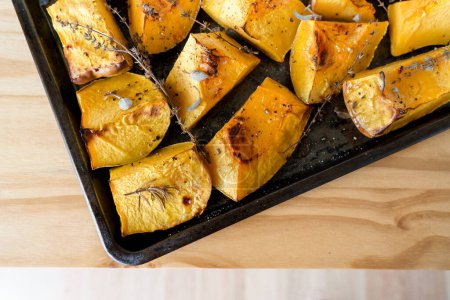 Pieces of organic oven roasted butternut squash on a black baking sheet pan with fresh thyme and sage herbs in natural light.