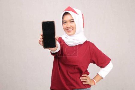 Téléchargez les photos : Jeune femme hijab en chemise rouge montrant téléphone portable écran noir à l'appareil photo avec belle expression de sourire, concept d'indépendance indonesia - en image libre de droit