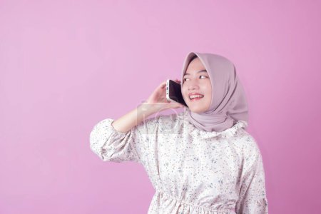 fun and happy indonesian hijab teen woman using mobile phone towards isolated pink studio empty room