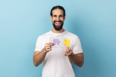 Photo for Portrait of man with beard wearing white T-shirt holding yellow and purple puzzle pieces, solving tasks, looking at camera with toothy smile. Indoor studio shot isolated on blue background. - Royalty Free Image
