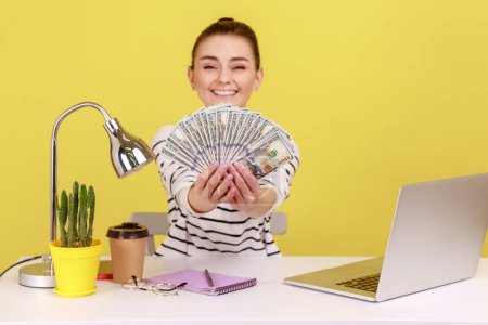 Photo for Delighted rich woman office manager holding hundred dollar bills in hand sitting at workplace, satisfied about big profit. Indoor studio studio shot isolated on yellow background. - Royalty Free Image