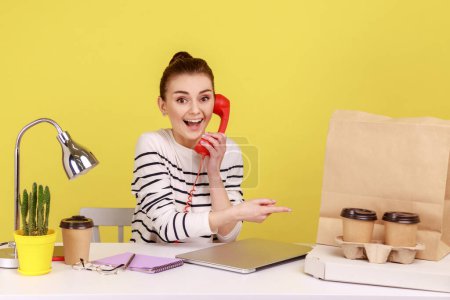 Photo for Happy woman pointing at pizza box cups of coffee and paper bag with purchase with smile and talking landline phone, enjoying fast food delivery. Indoor studio studio shot isolated on yellow background - Royalty Free Image