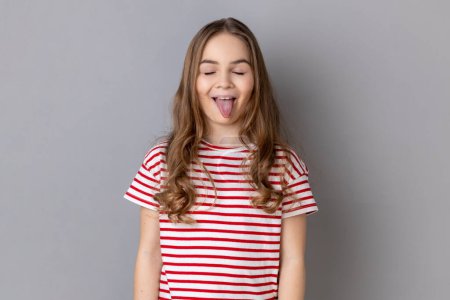 Porträt eines positiven, fröhlichen kleinen Mädchens mit gestreiftem T-Shirt, das mit geschlossenen Augen und herausgestreckter Zunge steht und kindliches Verhalten demonstriert. Indoor Studio isoliert auf grauem Hintergrund gedreht.