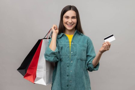 Photo for Happy satisfied woman holding and showing limitless credit card and shopping bags, happy with great shopping, final sale, wearing casual style jacket. Indoor studio shot isolated on gray background. - Royalty Free Image