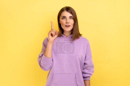 Photo for Portrait of inspired woman raising finger and having genius idea, looking amazed by sudden smart thought, wearing purple hoodie. Indoor studio shot isolated on yellow background. - Royalty Free Image