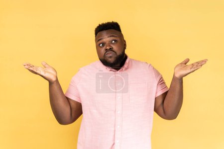 Photo for I dont know. Portrait of confused man wearing pink shirt shrugging shoulders as doesn't know answer, can`t make decision, being uncertain, not sure. Indoor studio shot isolated on yellow background. - Royalty Free Image