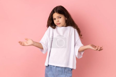 Photo for Portrait of confused little girl in white T-shirt shrugging shoulders with no idea gesture, clueless embarrassed face, don't know the correct answer. Indoor studio shot isolated on pink background. - Royalty Free Image