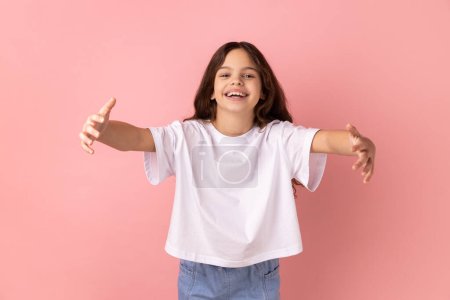 Free hugs, come into my arms. Portrait of little girl wearing white T-shirt stretching hands to camera and smiling broadly, going to embrace, share love. Indoor studio shot isolated on pink background