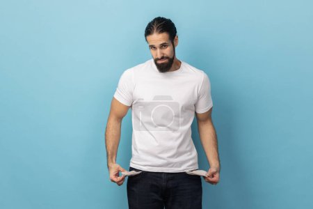 Photo for Portrait of man with beard wearing white T-shirt showing empty pockets and looking frustrated about loans and debts, has no money, jobless. Indoor studio shot isolated on blue background. - Royalty Free Image