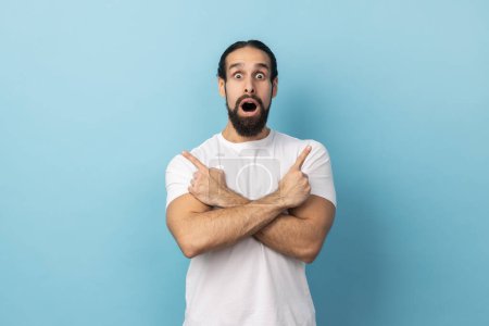Photo for Portrait of amazed surprised man with beard wearing white T-shirt standing with crossed hands and pointing to copy space on both sides. Indoor studio shot isolated on blue background. - Royalty Free Image