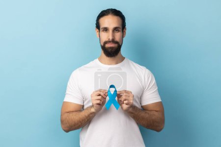 Photo for Portrait of serious responsible man with beard wearing white T-shirt holding blue awareness, disease symbol, looking at camera, support. Indoor studio shot isolated on blue background. - Royalty Free Image