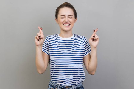 Photo for Portrait of hopeful beautiful young adult woman wearing striped T-shirt crossing fingers, hope for better, wishing for good luck, ritual. Indoor studio shot isolated on gray background. - Royalty Free Image