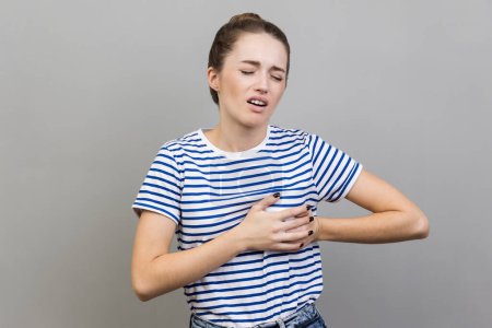 Photo for Acute pain in chest. Portrait of woman in striped T-shirt clutching breast and grimacing from painful cramp, heart attack at young age, cardiac disease. Indoor studio shot isolated on gray background. - Royalty Free Image