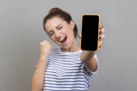Photo for Portrait of woman wearing striped T-shirt holding smartphone and smiling making yes gesture, celebrating online lottery or giveaway victory. Indoor studio shot isolated on gray background. - Royalty Free Image