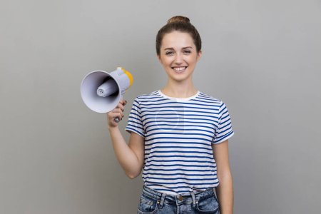 Photo for Portrait of woman wearing striped T-shirt having satisfied expression, holding megaphone in hands, looking at camera with toothy smile. Indoor studio shot isolated on gray background. - Royalty Free Image