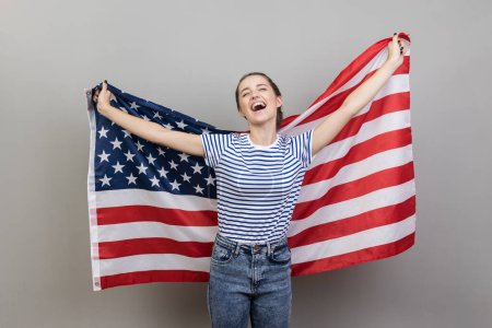 Photo for Portrait of cheerful young adult attractive woman wearing striped T-shirt holding big american flag, expressing happiness, screaming happily, rejoicing. Indoor studio shot isolated on gray background. - Royalty Free Image