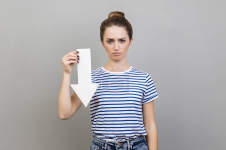 Photo for Portrait of sad upset woman wearing striped T-shirt showing white arrow pointing down, expressing sadness and sorrow, downgrade concept. Indoor studio shot isolated on gray background. - Royalty Free Image