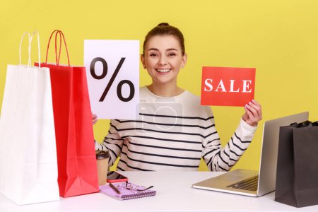Photo for Delighted woman holding sale card and paper with percent mark, announce discounts, sitting at workplace with laptop and paper shopping bags. Indoor studio studio shot isolated on yellow background. - Royalty Free Image