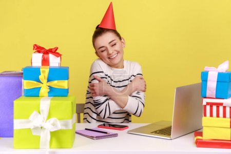Photo for Satisfied woman office employee with party cone sitting at workplace surrounded by many presents and embracing herself, celebrating birthday. Indoor studio studio shot isolated on yellow background. - Royalty Free Image