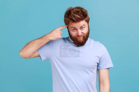 Téléchargez les photos : Portrait of young bearded man showing stupid gesture with finger near head, out of mind, accusing crazy dumb plan, reckless expression. Indoor studio shot isolated on blue background. - en image libre de droit