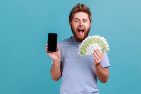 Téléchargez les photos : Portrait d'un homme barbu extrêmement heureux tenant des billets en euros et un téléphone portable avec écran vide, maquettes pour la publicité. Studio intérieur tourné isolé sur fond bleu. - en image libre de droit