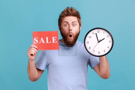 Photo for Portrait of shocked astonished handsome bearded man holding clock and card with sale inscription, announcing finishing huge discounts. Indoor studio shot isolated on blue background. - Royalty Free Image