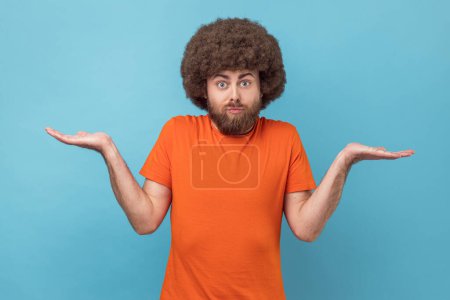 Photo for Clueless confused man with Afro hairstyle in T-shirt shrugging shoulders as doesn't know answer, can`t make decision, being uncertain, not sure. Indoor studio shot isolated on blue background. - Royalty Free Image