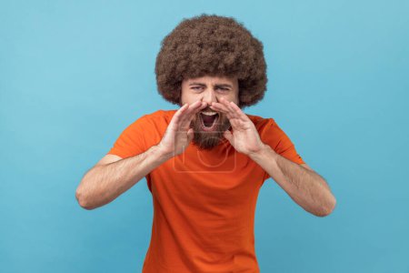Téléchargez les photos : Portrait d'un homme agressif en colère avec une coiffure afro portant un T-shirt orange debout, tenant les bras près de la bouche grande ouverte et criant. Studio intérieur tourné isolé sur fond bleu. - en image libre de droit
