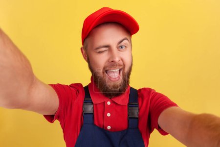 Photo for Playful delighted worker man wearing blue uniform taking selfie, looking at camera winking and showing tongue out, point of view of photo. Indoor studio shot isolated on yellow background. - Royalty Free Image