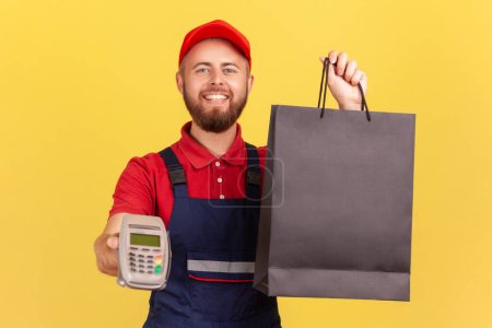 Photo for Portrait of bearded happy positive courier man bringing order in black shopping bag and giving pos terminal to client to pay for purchase. Indoor studio shot isolated on yellow background. - Royalty Free Image