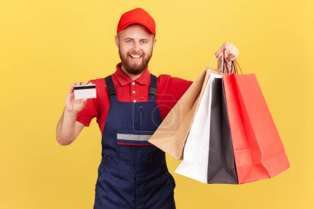 Photo for Portrait of positive smiling deliveryman in blue uniform, red t-shirt and hat holding shopping bag and credit card, cashless payments. Indoor studio shot isolated on yellow background. - Royalty Free Image