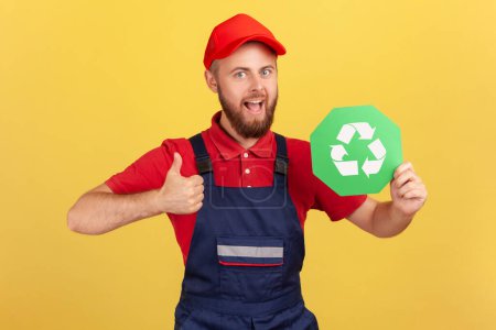 Photo for Portrait of satisfied worker man standing and holding recycling sign, thinking green, showing thumb up, wearing overalls and red cap. Indoor studio shot isolated on yellow background. - Royalty Free Image